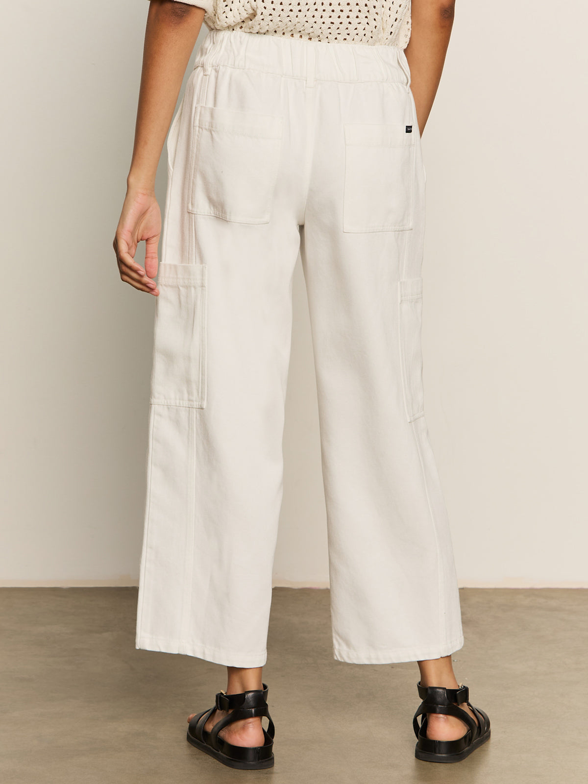 A person stands indoors, facing away, showing the lower back and legs in Sanctuary Clothing's area standard rise pant bleached white with patch pockets, paired with black sandals against a neutral background.