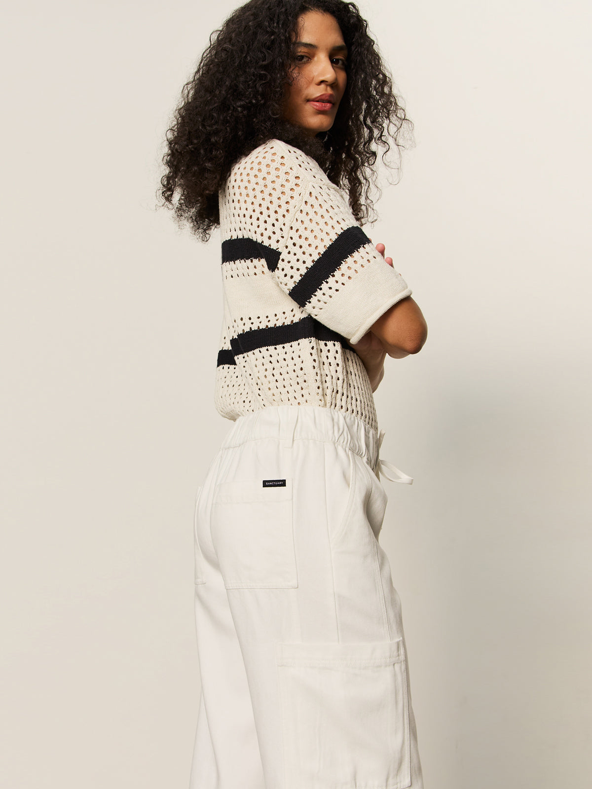 A person with long curly hair stands sideways in a white short-sleeve crochet top with black stripes and Sanctuary Clothing’s area standard rise pant in bleached white, featuring patch pockets, against a plain light background.