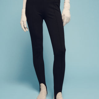 A person wearing a long-sleeved white top, stirrup legging black by Sanctuary Clothing, white gloves, and white socks stands against a light blue background. The image focuses on their legs and waist.