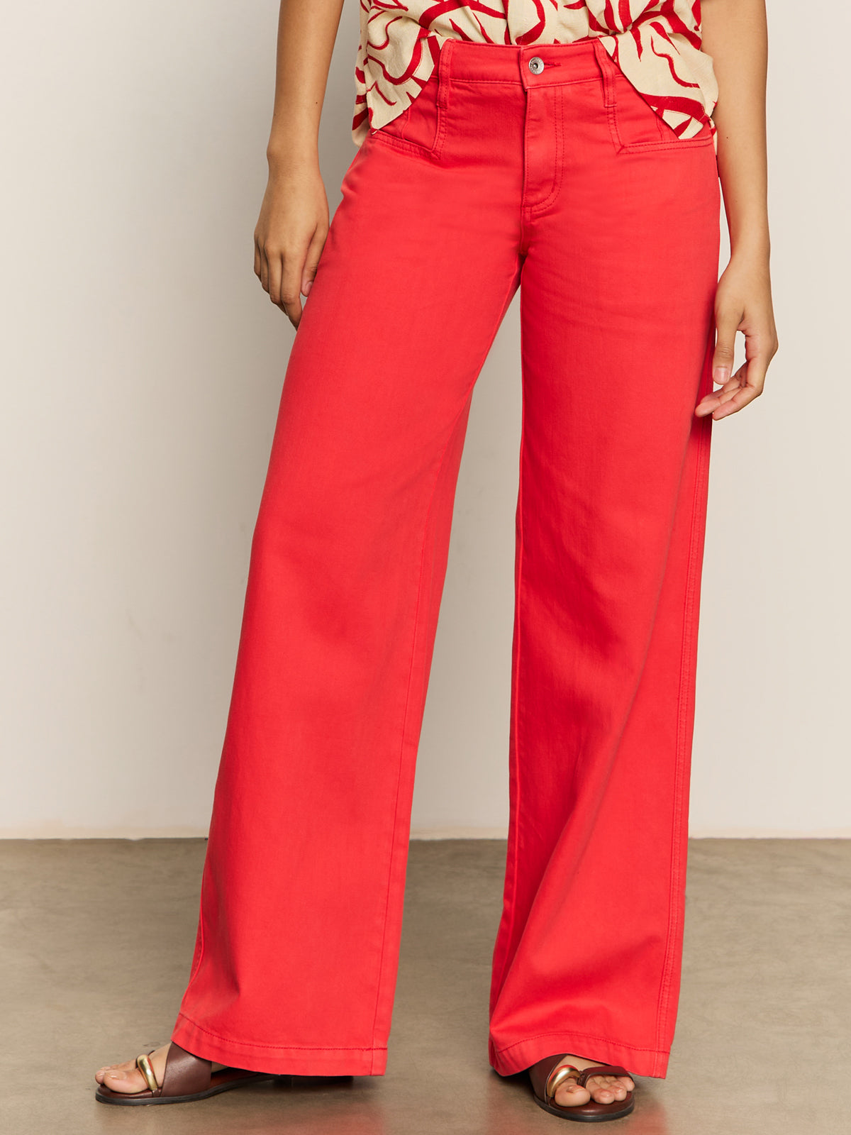 A person is casually standing, wearing Sanctuary Clothing's Highland Wide Leg Standard Rise Pant in cayenne with a patterned top. Their left hand rests on their side, and they wear open-toed sandals on a plain gray floor.
