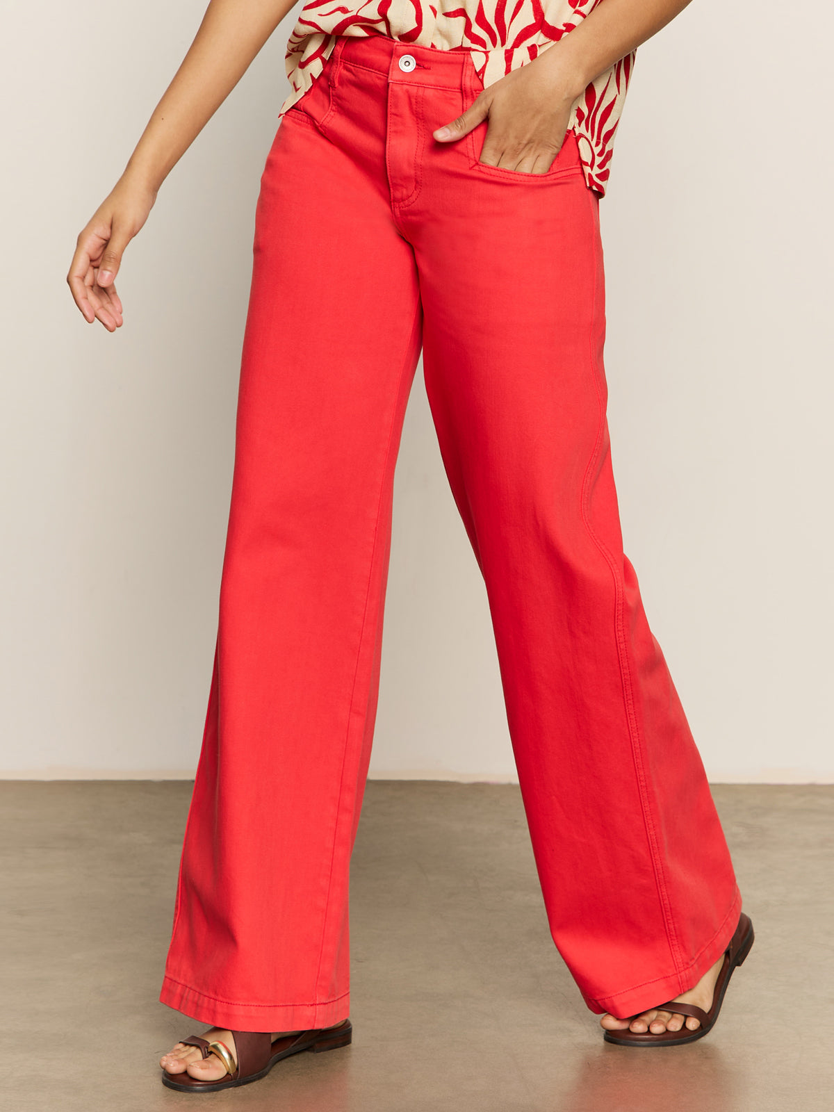 A person is standing barefoot on a concrete floor, wearing Sanctuary Clothing's highland wide leg standard rise pant in cayenne and a patterned top, with one hand in a pocket and their face not visible.