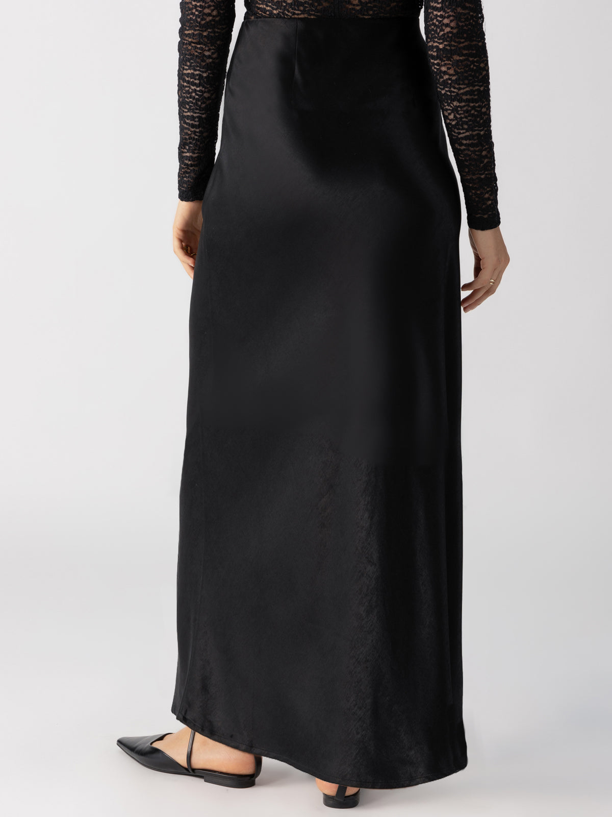 A person is standing with their back facing the camera, adorned in Sanctuary Clothing's "Everyday Maxi Skirt Black" and a black lace long-sleeve top, paired with black pointed-toe flat shoes. The backdrop is a simple white.