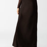 A person standing, wearing the Sanctuary Clothing Everyday Maxi Skirt in Coffee and beige ankle boots. They have on a brown patterned long-sleeve top. The background is plain white. The person's face is not visible in the image.