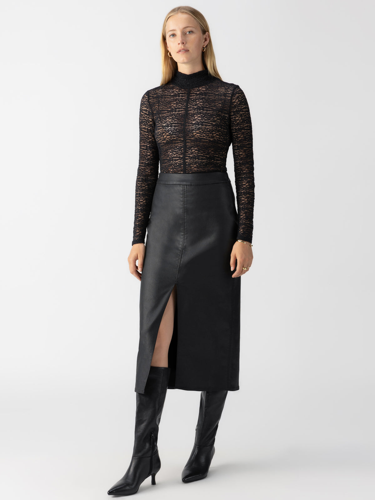 A person stands against a plain background, dressed in a black lace long-sleeve top paired with the Sanctuary Clothing Long Coated Skirt Black, which features a front slit. They also wear knee-high black leather boots. Their hair is down, and they have a neutral expression.