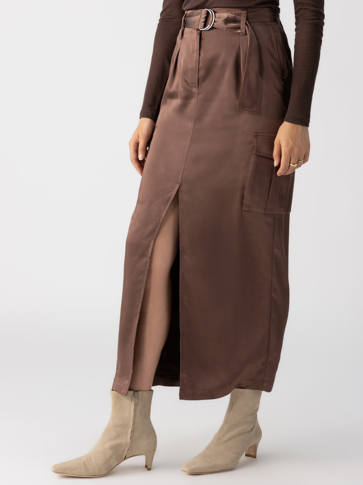 A person is wearing the Sanctuary Clothing Classy Cargo Skirt in Brown Sugar, which features a high waist, satin fabric, a front slit, and a belt. They have paired it with a long-sleeve dark brown top and light beige ankle boots with a low heel. Their right hand rests on their hip, and only the lower half of their body is visible.