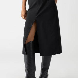 A person wearing the Classic Slit Skirt Black by Sanctuary Clothing, featuring a front slit and convenient pockets, paired with matching black knee-high boots. The individual is striking a pose with one hand tucked into a pocket, set against a plain white background.