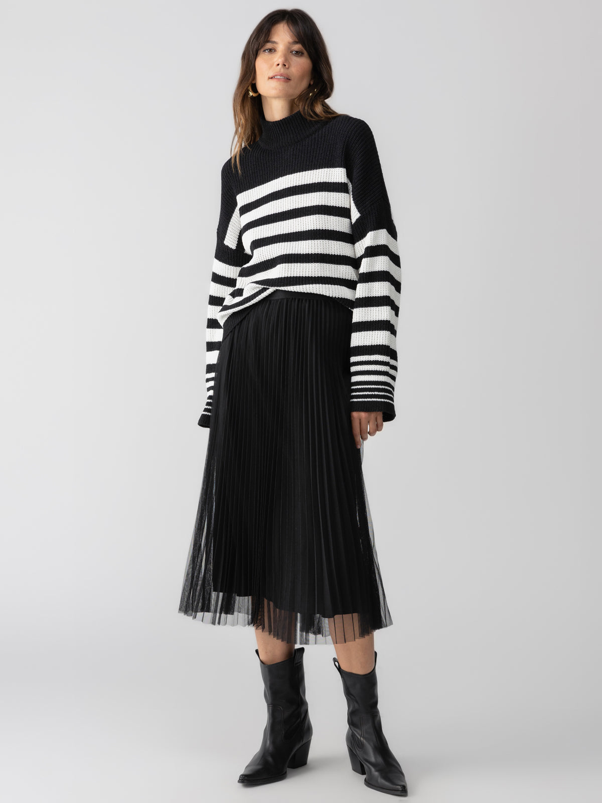 A woman stands against a plain background wearing a black and white striped sweater with long sleeves and the Tulle Pleated Midi Skirt in Black by Sanctuary Clothing. She has shoulder-length hair and is wearing black ankle boots.