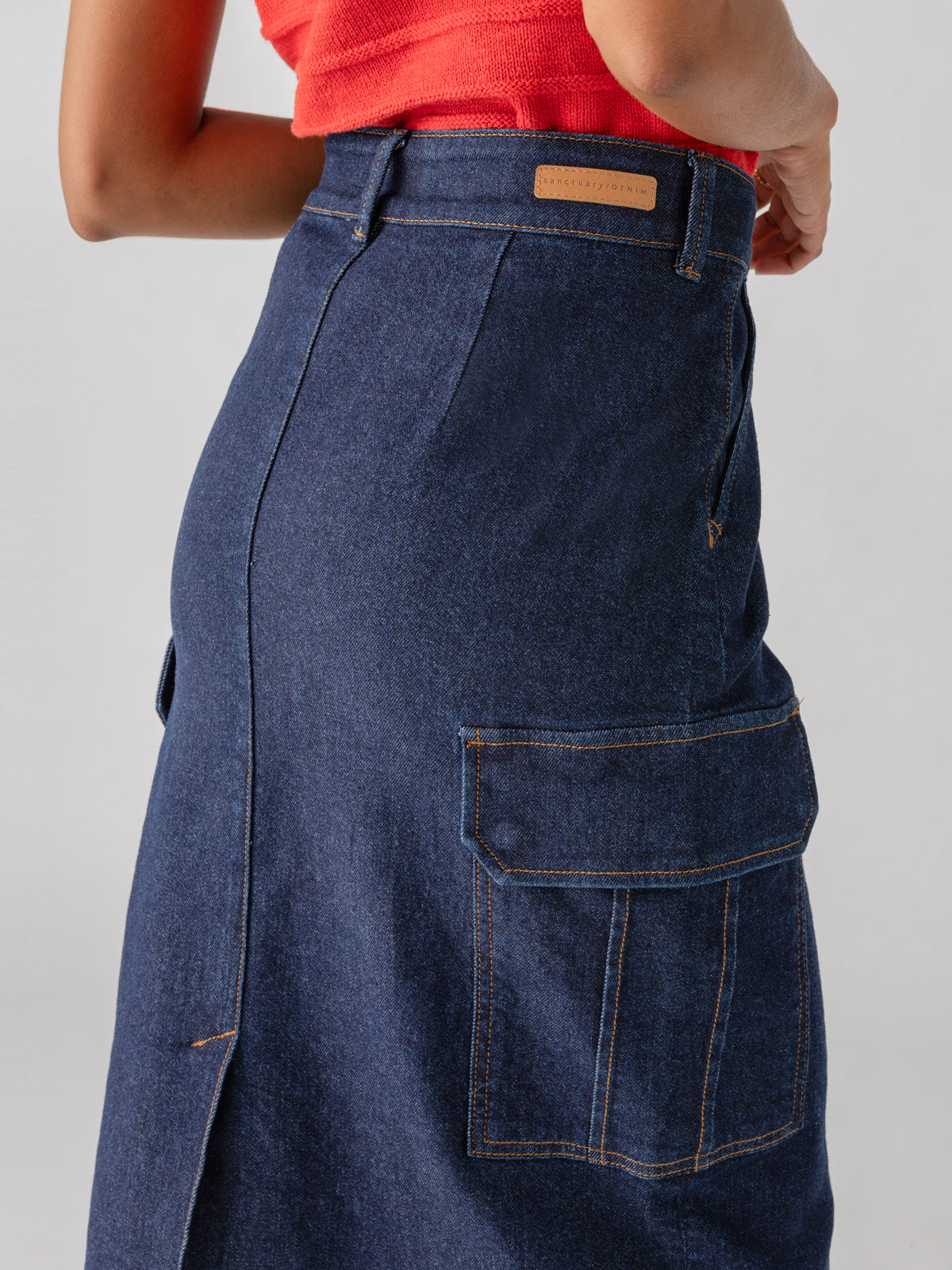 Image of a person wearing the Sanctuary Clothing Essential Cargo Skirt Magnetic in dark blue denim, featuring cargo pockets and a side vent, paired with a red sleeveless top. The shot is from the back, highlighting the skirt's design details.