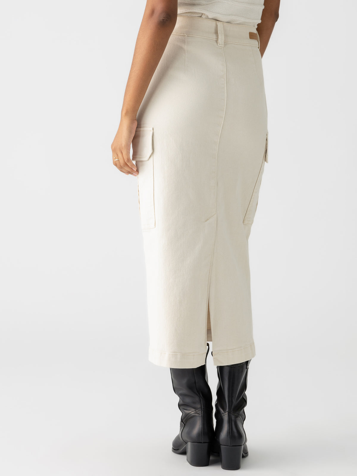 A person wearing the Essential Cargo Skirt in French Vanilla by Sanctuary Clothing, which features a high waist, midi length, utility pockets, and a back slit. They are also sporting black mid-calf heeled boots. The image is taken from behind against a plain white background.