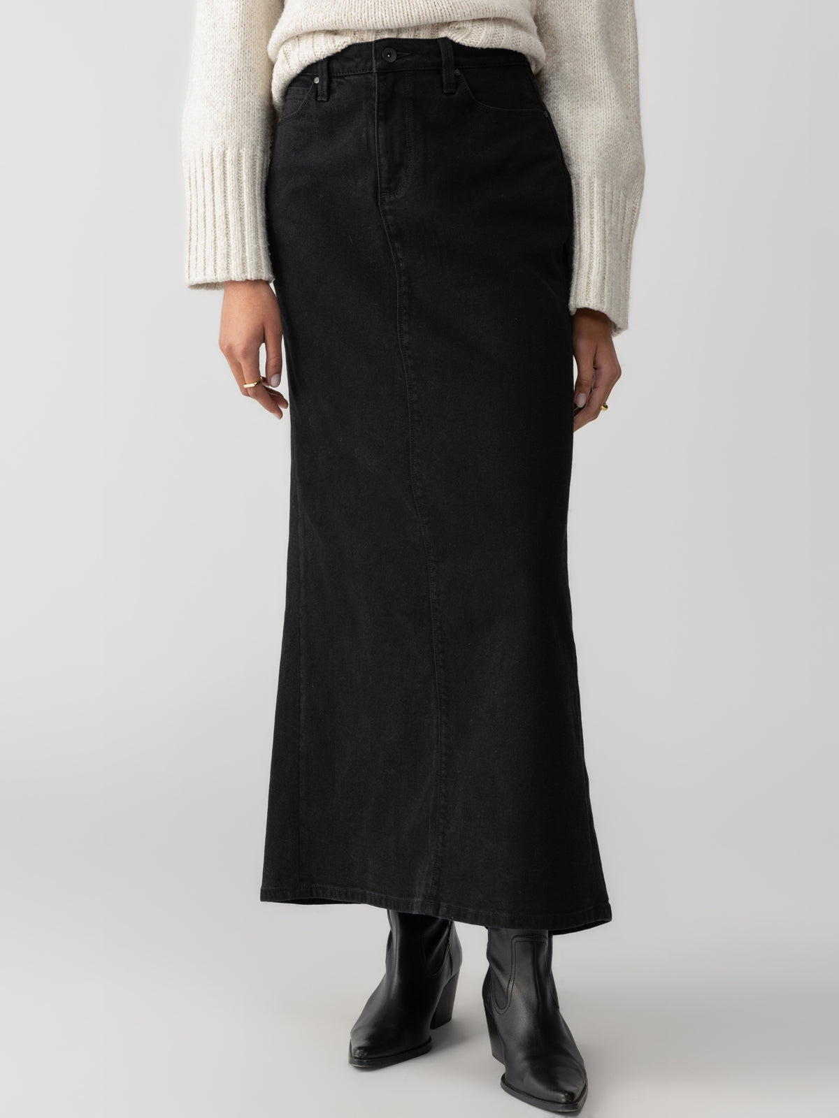 A person wearing a cream knit sweater, a long Denim Maxi Skirt Vintage Black by Sanctuary Clothing, and black boots stands against a plain white background. The image focuses on the lower half of their body, displaying the clothing and footwear.