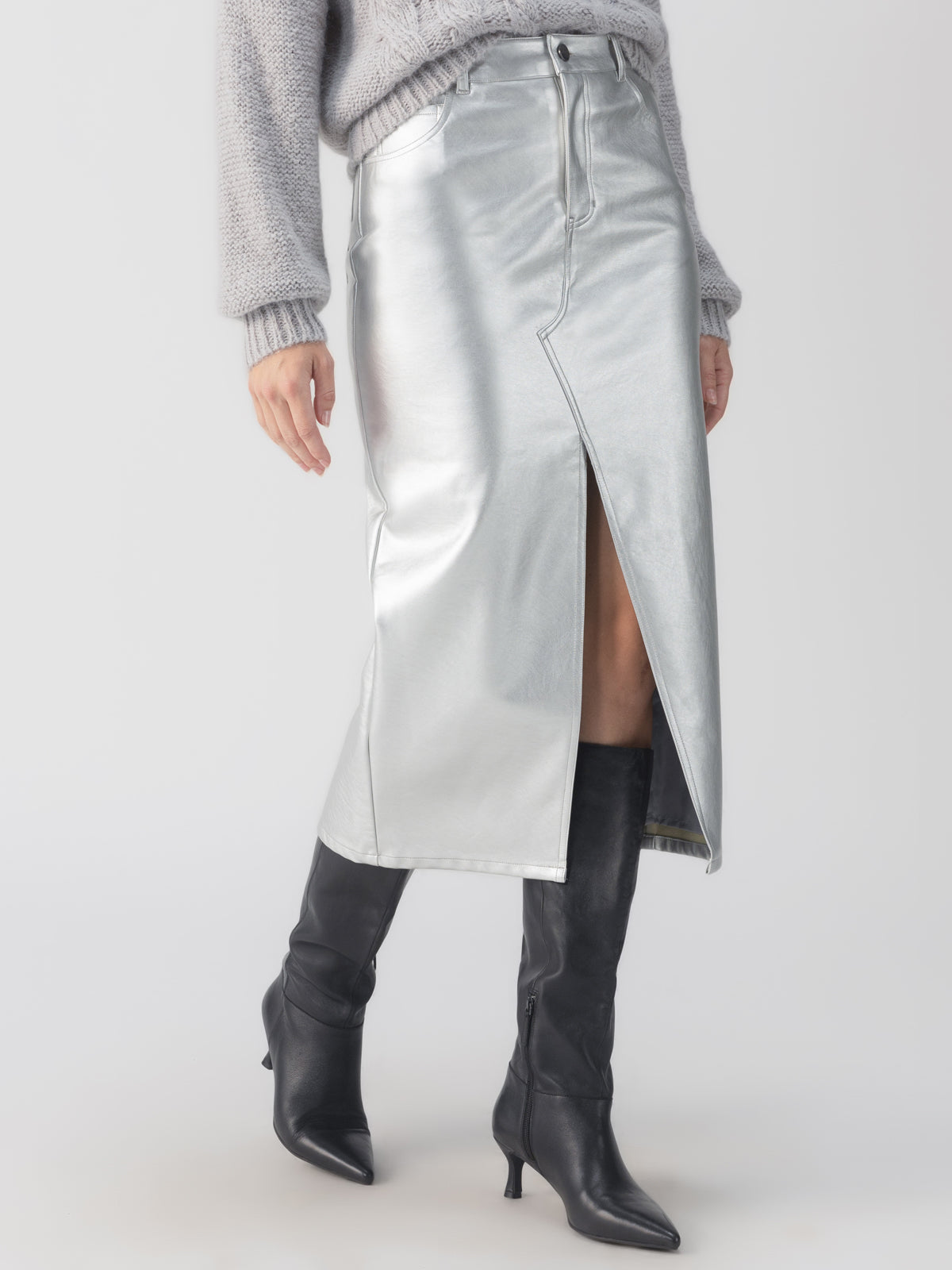 A person is wearing Sanctuary Clothing's "Leather Like Midi Skirt Silver," featuring a front slit, paired with a gray textured sweater and black knee-high boots. The outfit is styled against a plain white background.