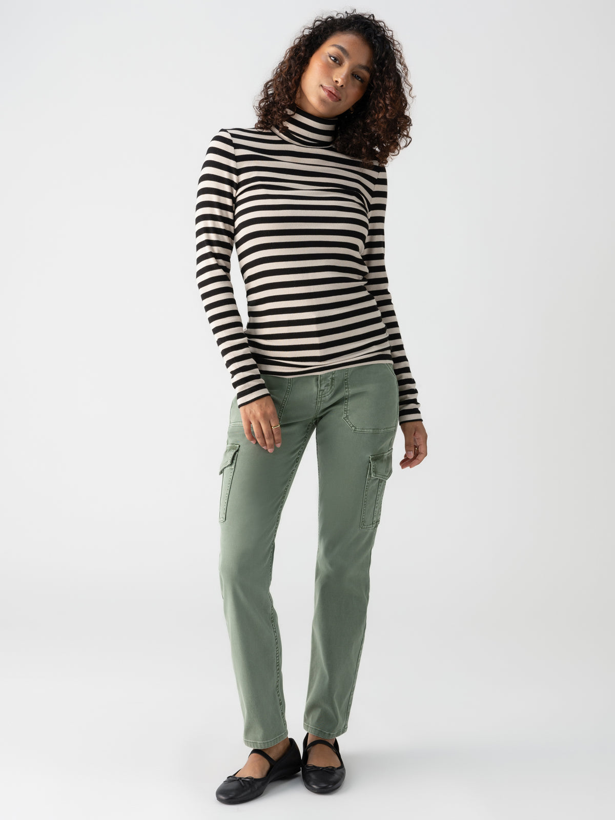 A woman stands against a plain white background, wearing the "Essential Turtleneck Toasted Almond Black Mini Stripe" by Sanctuary Clothing, paired with sage green cargo pants. She is also wearing black ballet flats and has curly shoulder-length hair. She looks to the front with a neutral expression.