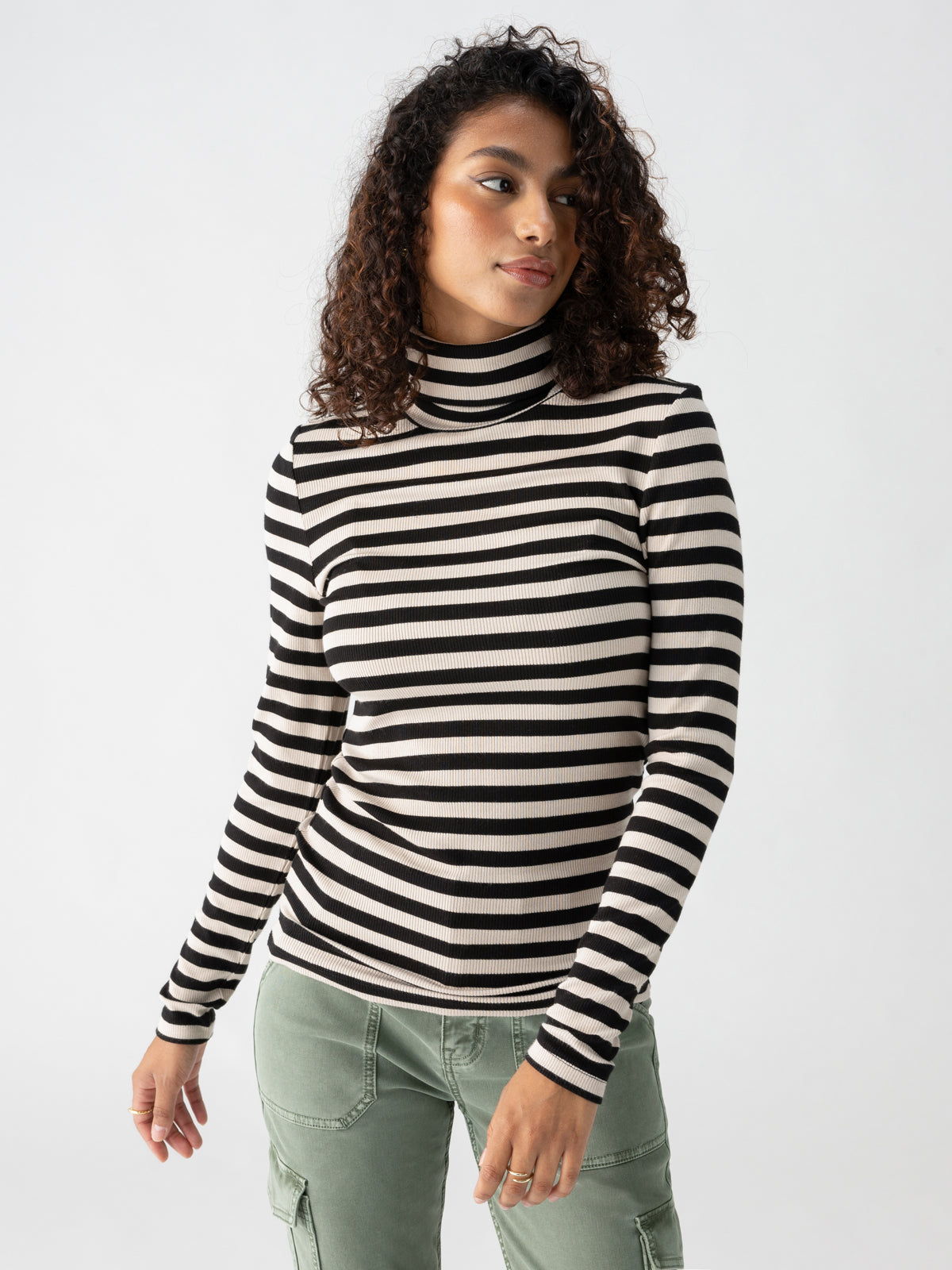 A person stands against a plain background wearing the Essential Turtleneck Toasted Almond Black Mini Stripe from Sanctuary Clothing and light green cargo pants. They have curly hair and are gazing slightly to their right with a neutral expression.