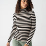 A person stands against a plain background wearing the Essential Turtleneck Toasted Almond Black Mini Stripe from Sanctuary Clothing and light green cargo pants. They have curly hair and are gazing slightly to their right with a neutral expression.