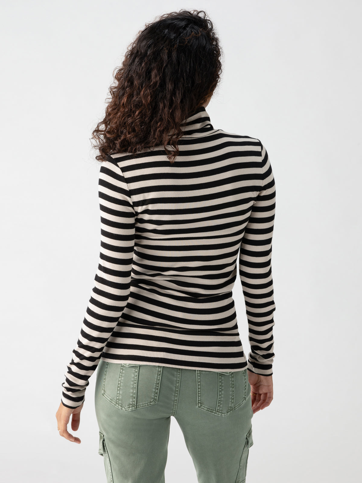 A person with long, curly hair stands facing away from the camera, wearing the Essential Turtleneck Toasted Almond Black Mini Stripe by Sanctuary Clothing and light green cargo pants. The background is plain and light-colored.