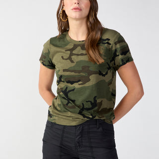 A person with long hair stands against a plain background, wearing Sanctuary Clothing's The Perfect Tee Hiker Camo and black cargo pants. They have their hands in their pockets, are looking directly at the camera, and are sporting hoop earrings.