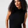 A woman with curly dark hair is wearing the Sanctuary Clothing Essential Sleeveless Mock Neck Top in black, paired with olive-green pants. She is standing against a plain, light gray background, looking towards the camera with a neutral expression.