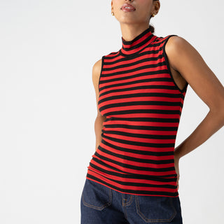 A person stands confidently against a plain background, wearing the Essential Sleeveless Mock Neck Black Cherry Red Stripe top from Sanctuary Clothing and dark blue jeans. They have one hand on their hip, and their hair is pulled back. The expression on their face is neutral.