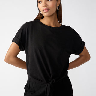 A woman poses confidently against a plain background. She wears the All Day Tie Tee Black from Sanctuary Clothing's Inclusive Collection, paired with black pants. Her straight hair is parted to one side, and she accessorizes with hoop earrings. With her hands resting on her hips, she gazes slightly to the side.