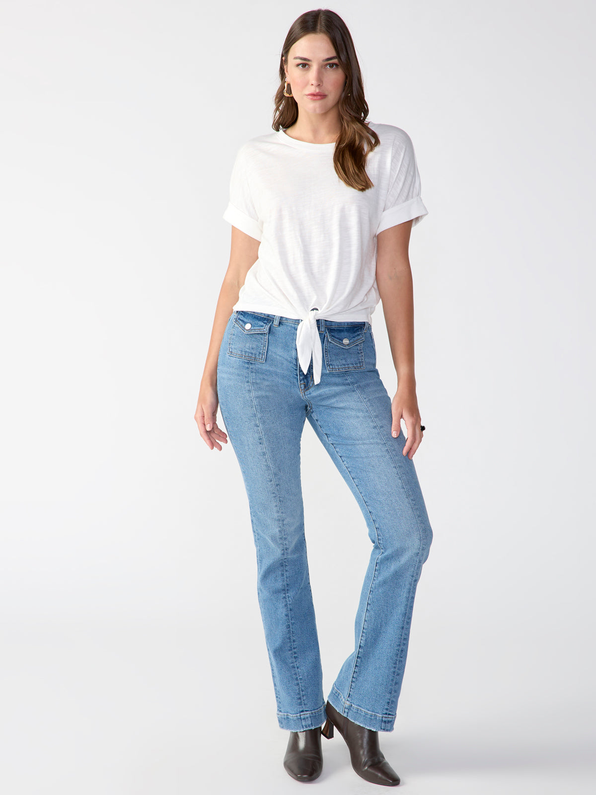 A woman with long hair is standing against a plain background. She is wearing the Sanctuary Clothing All Day Tie Tee White from the Inclusive Collection, paired with blue high-waisted bootcut jeans. She is also sporting dark boots. Her left hand rests on her hip, while her right arm hangs by her side.