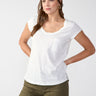 A woman with long, straight hair, partially tied back, stands against a plain background. She is wearing the Traveler Twist Tee White by Sanctuary Clothing and olive green cargo pants. Her hands are positioned on her hips.