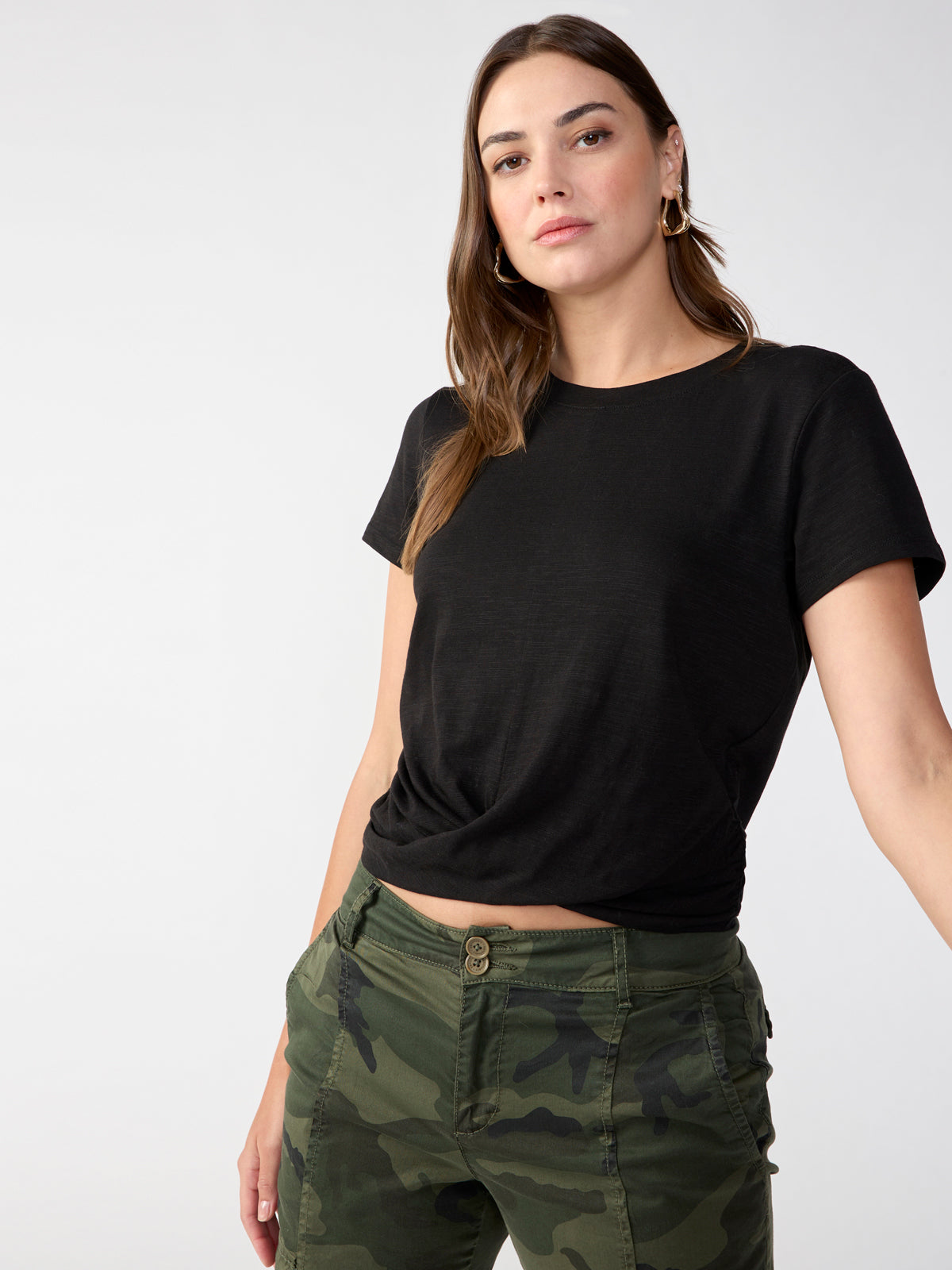 A person with shoulder-length brown hair is wearing a Highstreet Tee Black by Sanctuary Clothing and camouflage-patterned pants. They have a neutral expression and are standing against a plain, light-colored background.