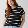 A woman with long, red hair is standing against a plain background, dressed in the Sanctuary Clothing's The Perfect Tee Black Stripe from the Inclusive Collection and light blue pants. She is smiling slightly with her hands in her pockets and accessorized with a gold necklace.