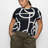 A woman wearing "the perfect tee graphic lines black inclusive collection" by Sanctuary Clothing and olive green pants stands with her hands in her pockets, looking down against a plain white background.