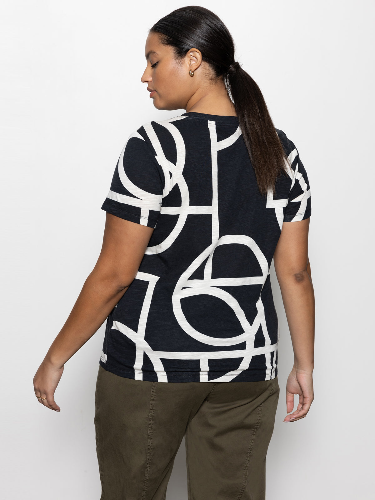 A person with long hair is standing with their back to the camera, dressed in Sanctuary Clothing's "the perfect tee graphic lines black" from the inclusive collection, paired with green pants. They are set against a plain white background.