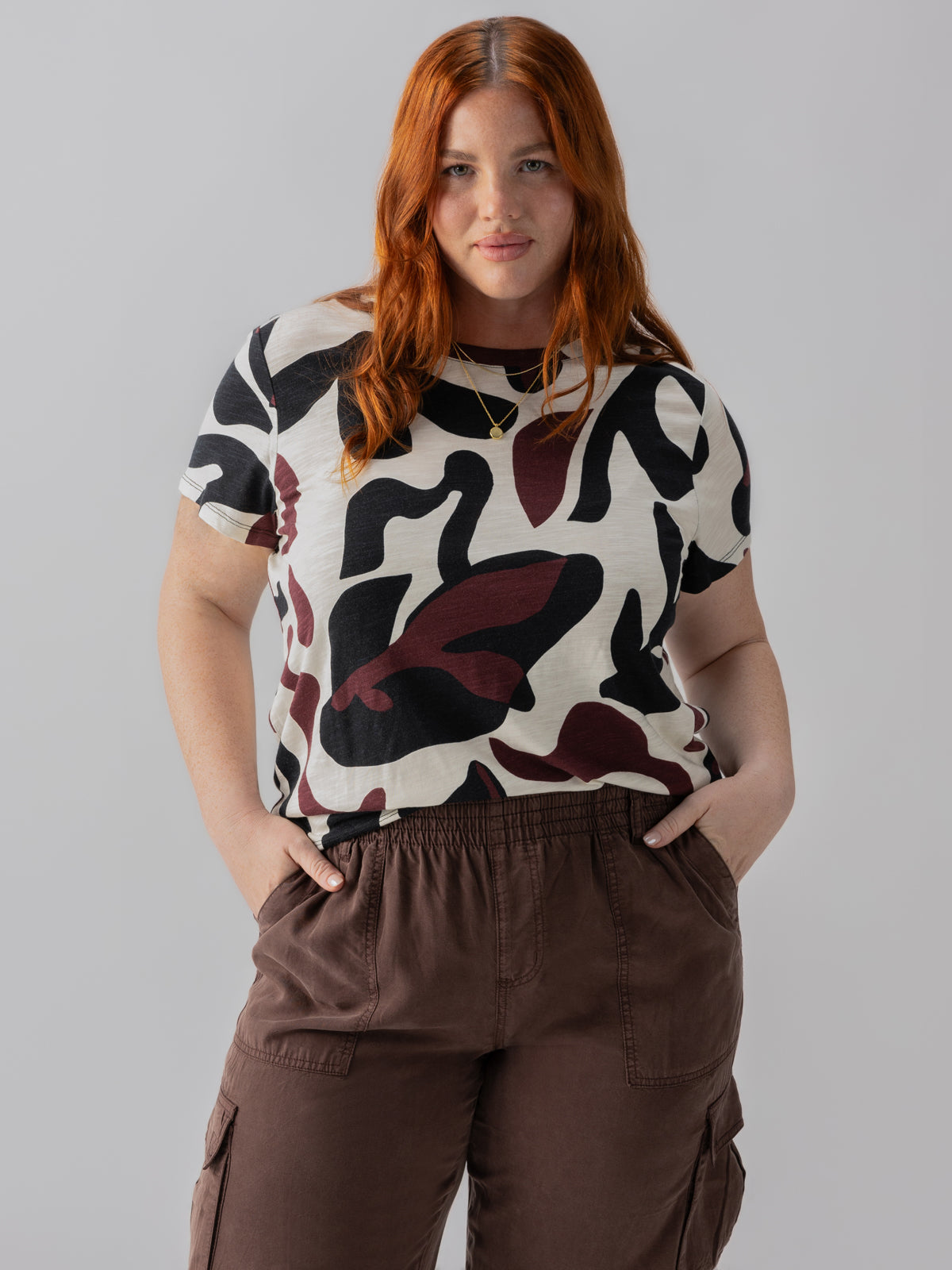 A person with long red hair is wearing The Perfect Tee Light Mineral Inclusive Collection from Sanctuary Clothing, paired with brown cargo pants. They have their hands in their pockets and are standing against a plain gray background.