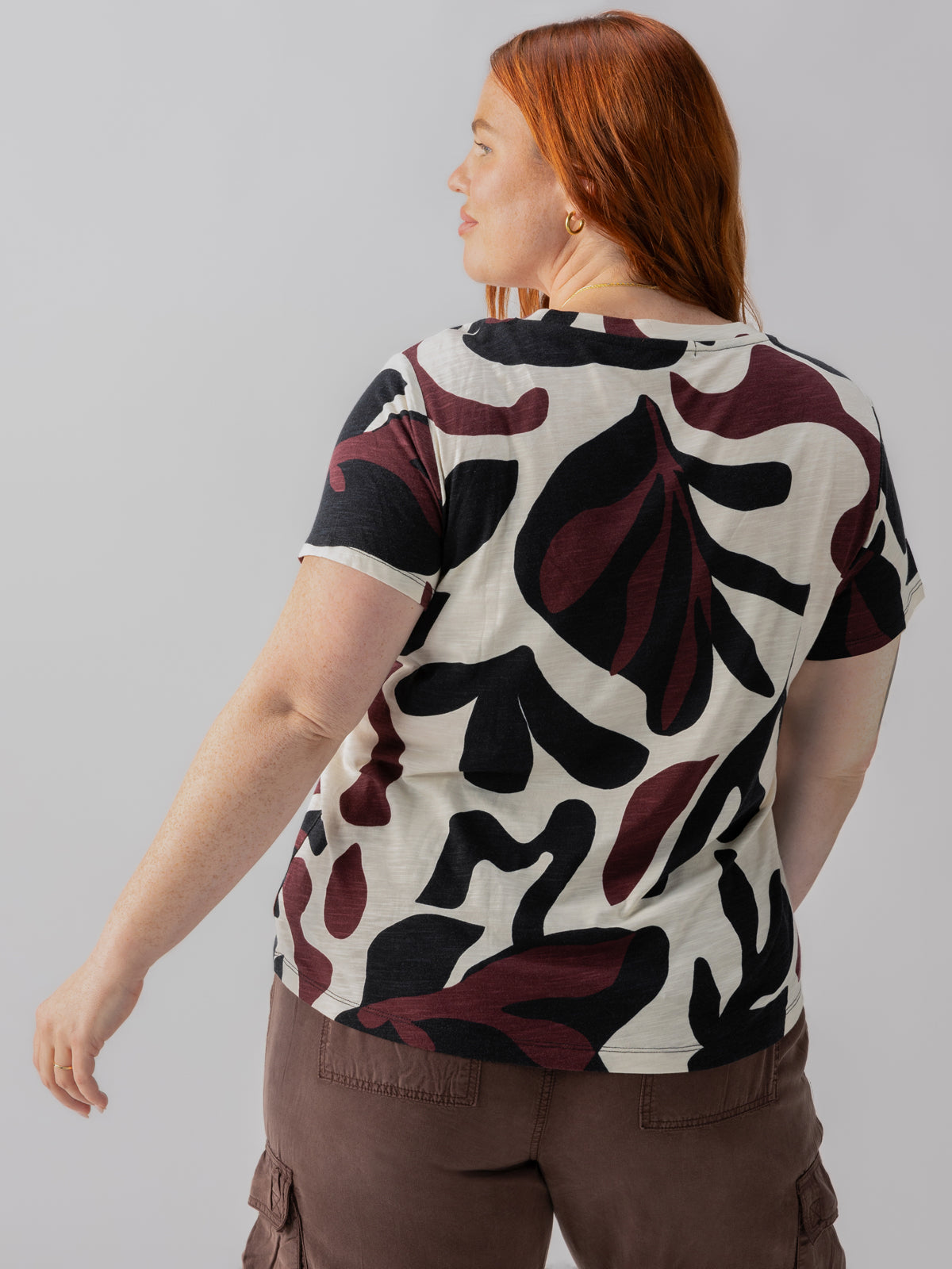 A person with long red hair is seen from behind, wearing The Perfect Tee Light Mineral Inclusive Collection from Sanctuary Clothing, which features a large abstract leaf pattern in shades of black, maroon, and beige. The person is also wearing brown cargo pants and has their right arm slightly bent.