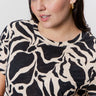 A person with dark hair wears gold hoop earrings and the Sanctuary Clothing "the perfect tee mirage extended sizing," a black and beige patterned shirt. They look slightly to the side with a neutral expression against a plain white background.