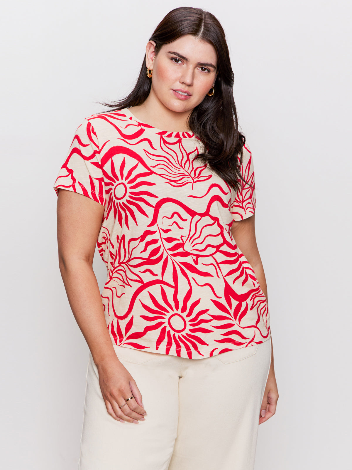 A woman models the Sanctuary Clothing "perfect tee sunny riviera extended sizing," featuring a bold red sun and wave pattern, paired with cream pants against a plain white backdrop. She has long, dark hair.