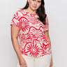A woman models the Sanctuary Clothing "perfect tee sunny riviera extended sizing," featuring a bold red sun and wave pattern, paired with cream pants against a plain white backdrop. She has long, dark hair.