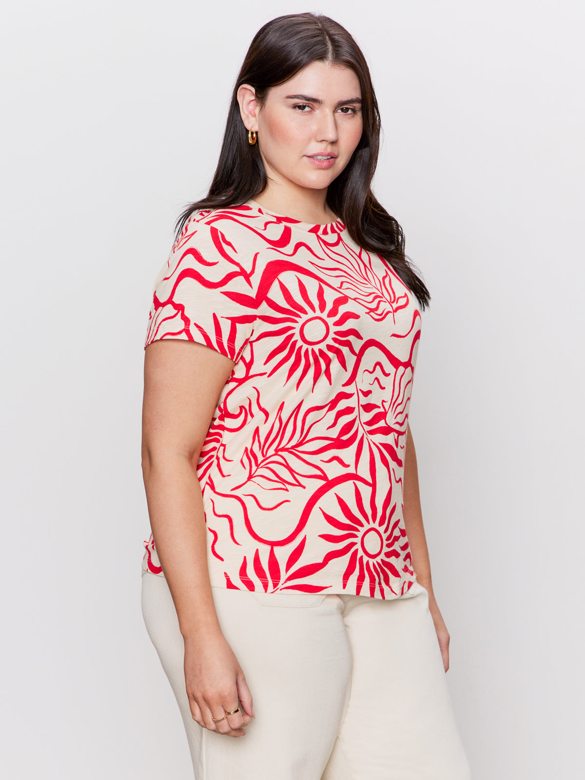 A woman wearing the Sanctuary Clothing's "The Perfect Tee - Sunny Riviera" with bold red sun and leaf patterns, confidently poses against a plain backdrop. She complements the look with light-colored pants.