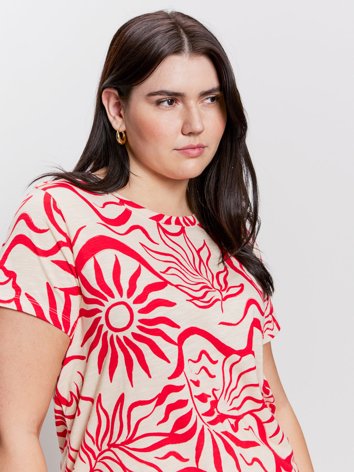 A person with long dark hair dons Sanctuary Clothing's "the perfect tee sunny riviera extended sizing," featuring a cream background with red abstract sun designs. Complementing gold hoop earrings, they gaze sideways against a plain backdrop.