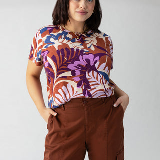 A person with shoulder-length dark hair stands confidently with hands in the pockets of their brown cargo pants. They are wearing Sanctuary Clothing's "The Perfect Tee South Palm Inclusive Collection," a colorful shirt featuring a bold tropical leaf pattern in shades of purple, white, and orange. The background is plain light gray.