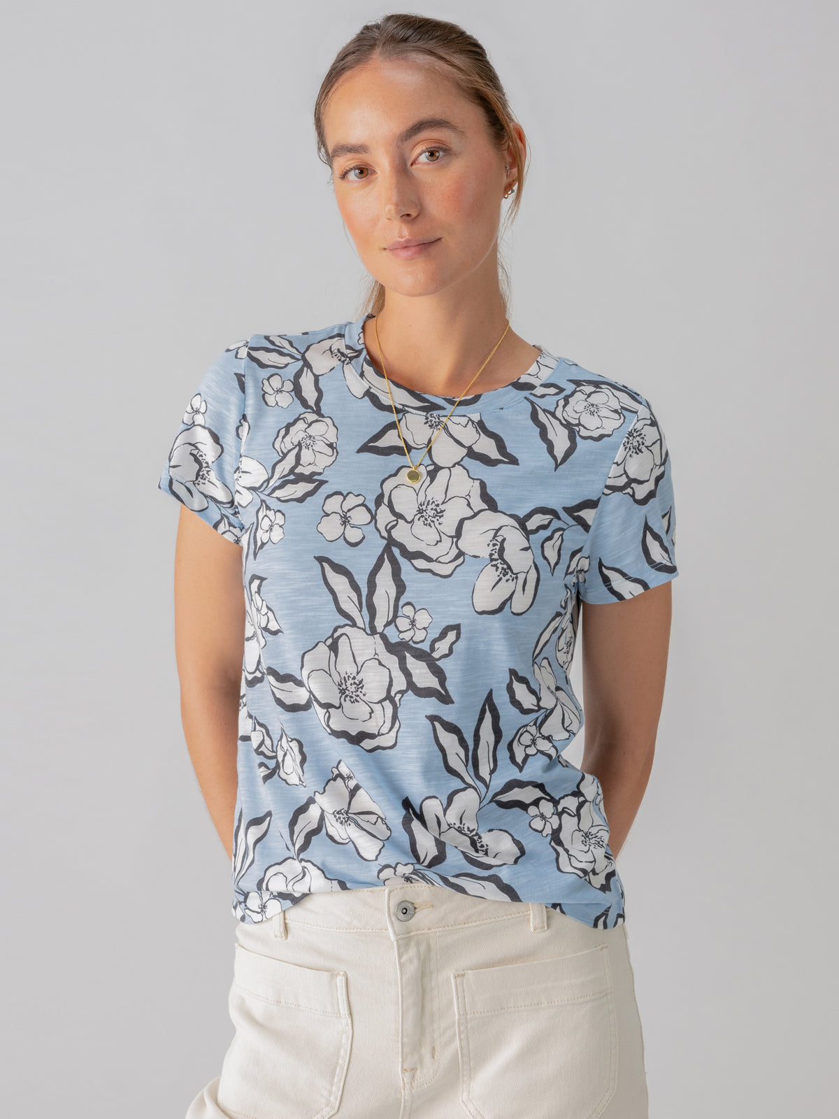 A woman with light brown hair tied back is wearing The Perfect Tee Blue Bliss Floral by Sanctuary Clothing and beige pants. She is standing against a plain background, looking at the camera with a slight smile. She has a gold pendant necklace.