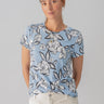 A woman with light brown hair tied back is wearing The Perfect Tee Blue Bliss Floral by Sanctuary Clothing and beige pants. She is standing against a plain background, looking at the camera with a slight smile. She has a gold pendant necklace.