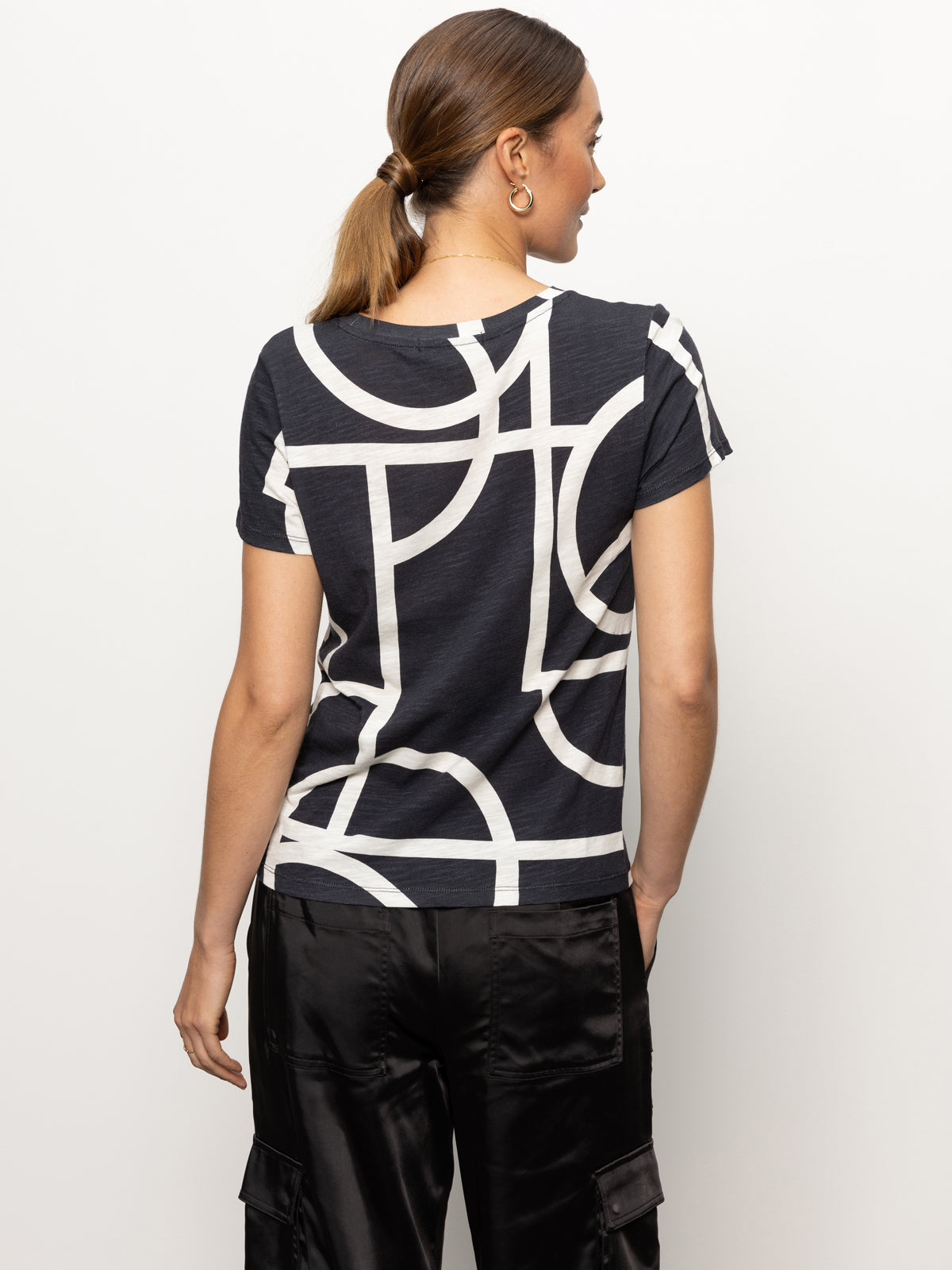 A person with long hair tied in a ponytail is wearing "the perfect tee graphic lines black" by Sanctuary Clothing, paired with sleek black satin pants. They are facing away, highlighting the distinctive geometric pattern on the back of the shirt against a plain white background.