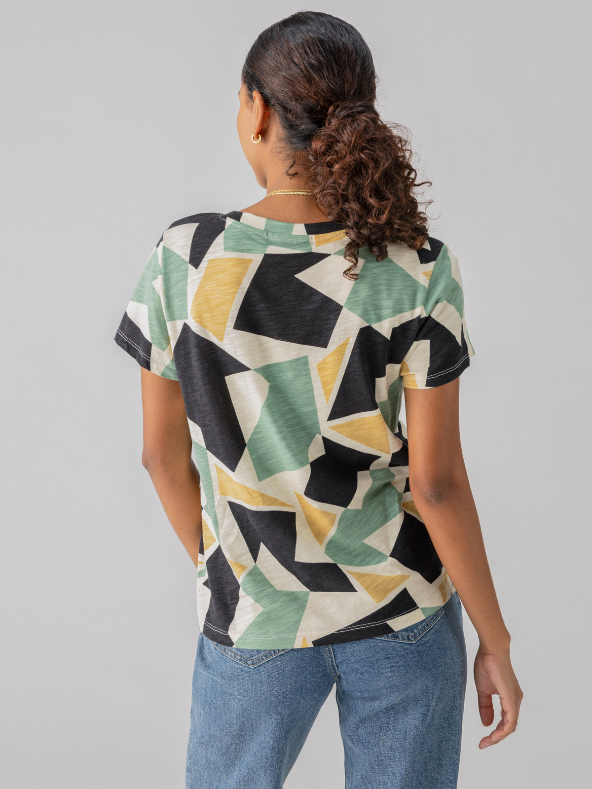 A person with curly hair is standing with their back to the camera. They are wearing The Perfect Tee Kaleidoscope by Sanctuary Clothing, featuring a short-sleeve design with a geometric pattern in green, beige, and black colors, paired with light blue jeans. The background is plain light gray.