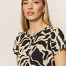 A person with long, straight hair wearing Sanctuary Clothing's "the perfect tee mirage," featuring a beige and black abstract pattern, looks calmly at the camera against a plain, light background.