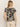 A person with long hair stands facing away, wearing Sanctuary Clothing's "The Perfect Tee Mirage" in a beige and black animal print, paired with beige shorts against a light gray background.