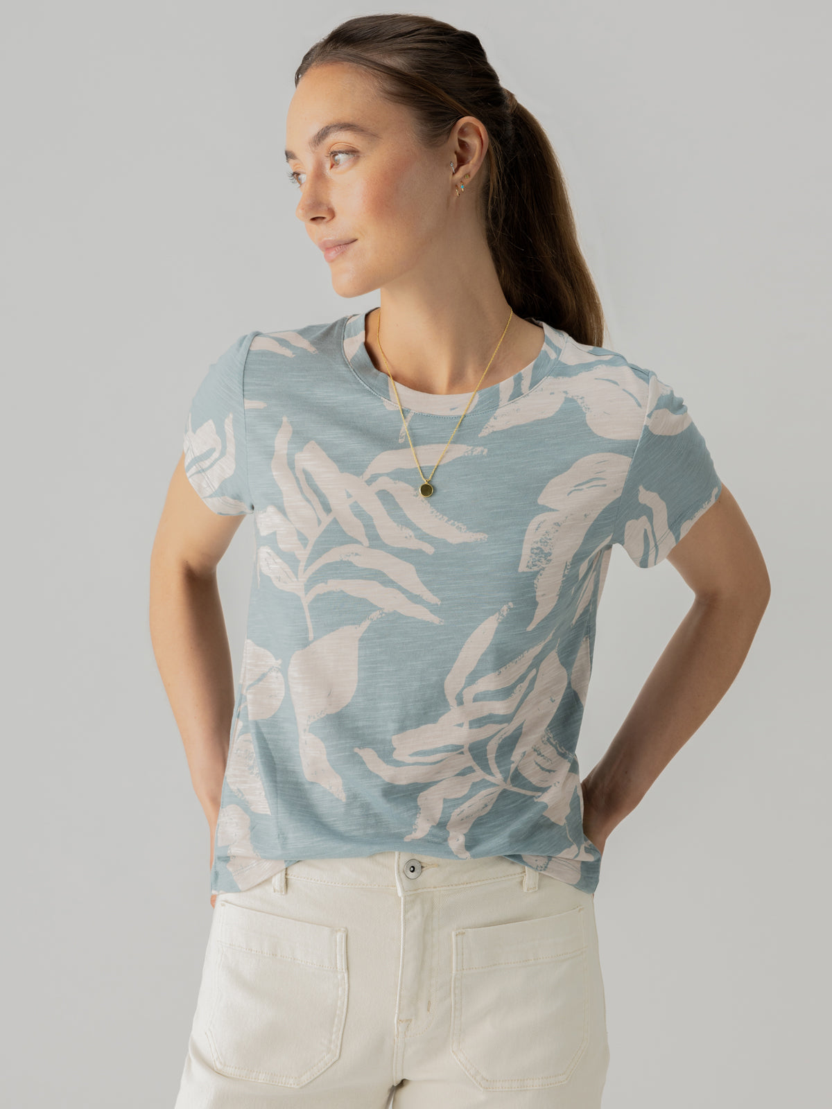 A woman wearing Sanctuary Clothing's The Perfect Tee Shadow Palms, a blue shirt with a light floral pattern, stands against a plain background. She has her long hair tied back and is wearing white pants, a necklace, and small earrings. She is looking to her left with her hands resting on her hips.