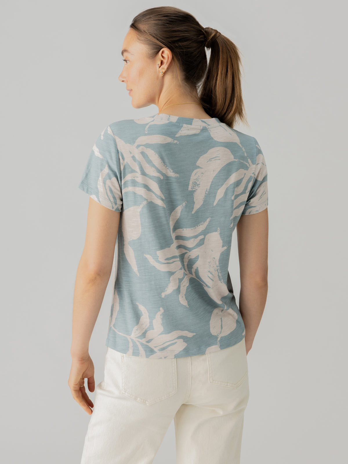 A woman stands with her back to us, looking to her left. She is wearing "The Perfect Tee Shadow Palms" from Sanctuary Clothing, a light blue t-shirt featuring a white abstract print, paired with white pants. Her hair is tied back in a ponytail, and she is positioned against a plain, light-colored background.