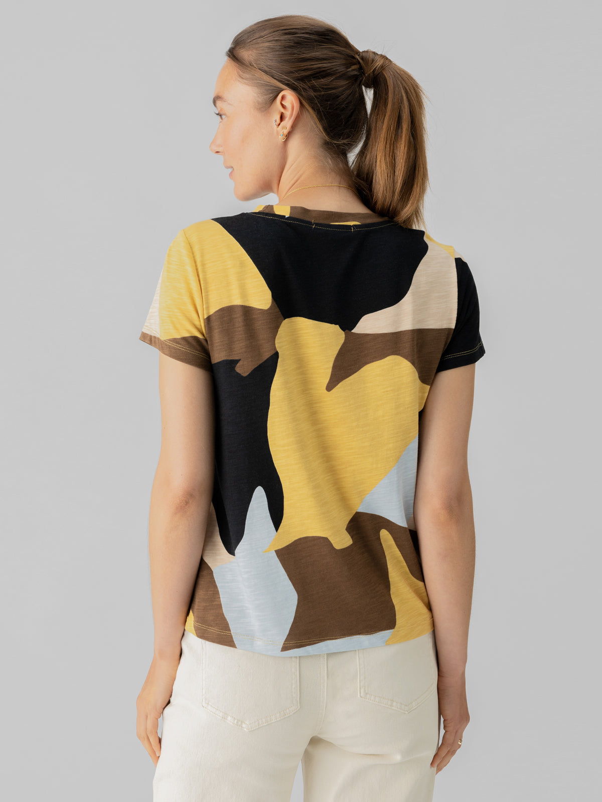 A person with a ponytail stands facing away, wearing Sanctuary Clothing's short-sleeved "The Perfect Tee Sky Escape," which features large, abstract shapes in yellow, black, brown, and light blue. They are also wearing light-colored pants against a plain gray background.