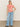 A long-haired person stands against a neutral background, wearing Sanctuary Clothing's "The Perfect Tee Sunny Riviera," paired with wide-legged blue jeans. Their left hand is on their hip, and they sport black open-toed sandals.
