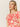 Someone with long blond hair is wearing "the perfect tee sunny riviera" from Sanctuary Clothing, which features bold red floral and abstract patterns on a beige background. They are gazing thoughtfully to the side against a plain backdrop.