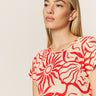 Someone with long blond hair is wearing "the perfect tee sunny riviera" from Sanctuary Clothing, which features bold red floral and abstract patterns on a beige background. They are gazing thoughtfully to the side against a plain backdrop.