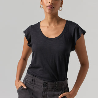 A person with short, slicked-back hair is facing the camera, wearing Sanctuary Clothing's black West Side Tee and dark high-waisted shorts. They have gold hoop earrings and their hands are in their shorts' pockets, standing against a plain gray background.